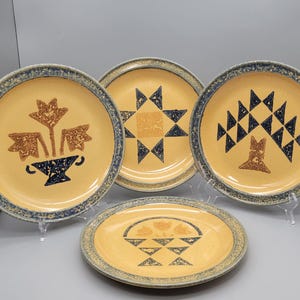 May include: Four ceramic plates with a yellow background and blue rims. Each plate features a different design in brown and navy blue: a floral arrangement, a star, a tree, and a basket. The plates are displayed on clear stands.