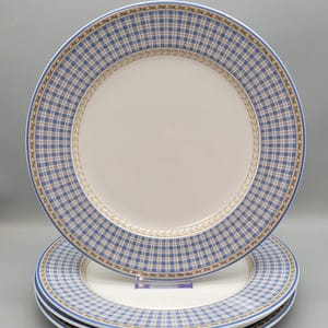 May include: A stack of three round ceramic dinner plates featuring a solid cream-colored center and a wide rim decorated with a blue and white plaid pattern, accented by a thin tan border with a repeating leaf-like motif along the inner edge.