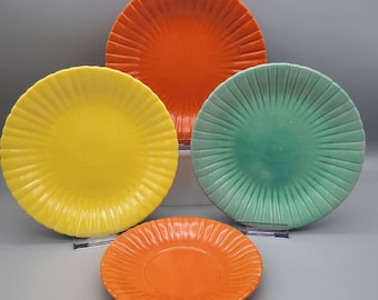 May include: Four colorful ceramic plates in shades of yellow, orange, and teal. Each plate has a ridged, radial design. The plates vary in size, with the largest in the background and the smallest in the foreground.