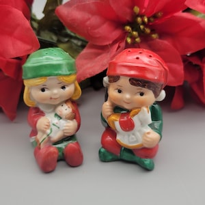 May include: Two ceramic Christmas elf figurines, one holding a doll and the other a rocking horse. They wear red, green, and white outfits with matching hats. Red poinsettia flowers are in the background.