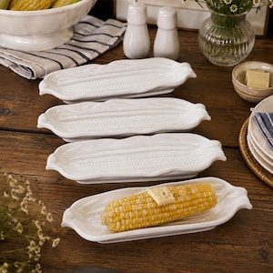 May include: A set of four white ceramic corn-shaped serving dishes, each featuring a textured surface mimicking corn husks. One dish holds a single cooked ear of yellow corn topped with a small square of butter, while the others are empty.