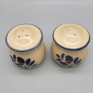 May include: A pair of ceramic salt and pepper shakers. The shakers are beige with a dark blue floral design and a dark blue rim. Each shaker has three holes on the top for dispensing.