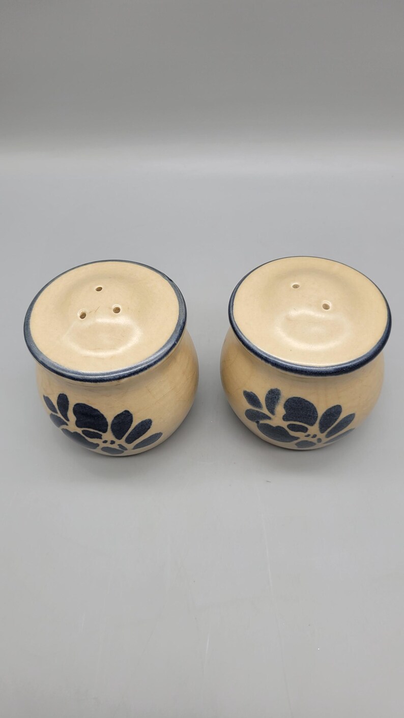 May include: A pair of ceramic salt and pepper shakers. The shakers are beige with a dark blue floral design and a dark blue rim. Each shaker has three holes on the top for dispensing.
