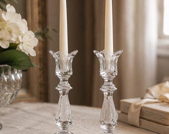 Puede incluir: Dos portavelas de cristal transparente con base facetada y parte superior en forma de tulipán, cada uno con una vela blanca encendida. El cristal presenta un patrón de corte reflectante que capta la luz, creando un aspecto brillante.