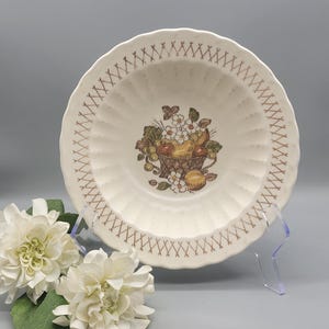 May include: Off-white ceramic bowl with a fruit basket design in the center. The bowl has a scalloped edge and a brown crosshatch pattern around the rim. The bowl is on a clear stand with white flowers in the foreground.