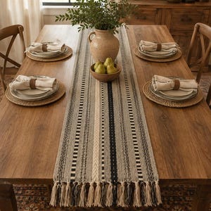May include: A rectangular woven table runner featuring a striped pattern in shades of beige, gray, and black with knotted fringe at both ends. The runner is centered on a wooden dining table with four place settings, each including a round placemat.