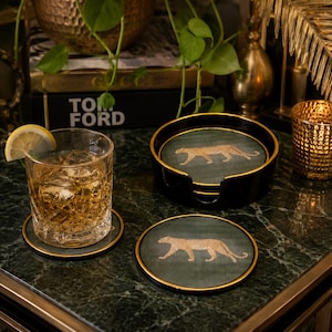 May include: A set of round coasters featuring a gold leopard illustration on a dark green background with gold-toned rims. One coaster sits under a glass of amber liquid with a lemon slice, while others are stored in a black circular holder.