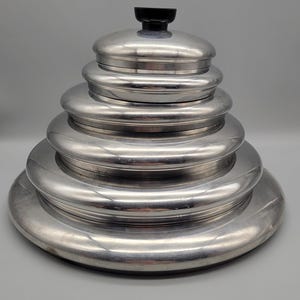 May include: A stack of polished aluminum serving bowls and lids, arranged in descending order by size. The top lid has a black handle. The bowls have a shiny, reflective surface, and the set is against a plain gray background.