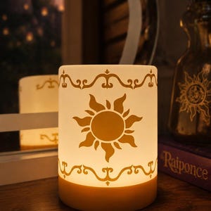 Puede incluir: Una lámpara cilíndrica blanca con un brillo interno cálido, que presenta un emblema de sol dorado en el centro y bordes decorativos de filigrana en la parte superior e inferior. La lámpara emite una luz suave que resalta el diseño del sol.