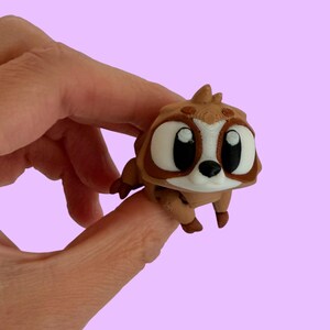 May include: A small, brown, three-dimensional printed figurine of a cartoon-style creature with large, white, oval-shaped eyes, black pupils, and a small black nose. The creature has a light-coloured face patch, pointed ears, and small limbs.