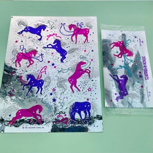 以下が含まれることがあります： 虹色のたてがみと尻尾を持つユニコーンが描かれた銀色のホログラフィックステッカーのシート。ステッカーはグリッドパターンで配置されており、キラキラとした仕上げになっています。シートには「Stick-r-Treats」という文字が印刷されています。