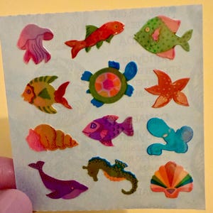May include: A sheet of colorful ocean-themed stickers featuring various sea creatures. The stickers include a jellyfish, fish, a turtle, a starfish, a seashell, a whale, a seahorse, and a clam shell. The stickers are in shades of pink, orange, green, blue, and purple.