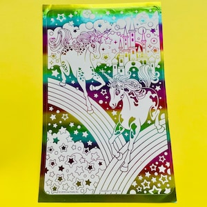 以下が含まれることがあります： 虹の上を走る2頭のユニコーンと、背景に城が描かれたホログラフィックステッカー。ステッカーは星で囲まれており、「COLGENO1170101F18」と「© 2018 Lisa Frank, Inc.」の文字が書かれている