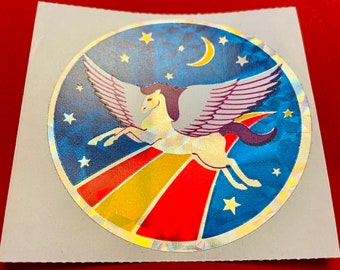 Puede incluir: Una pegatina holográfica con un caballo blanco alado volando sobre un arcoíris contra un cielo nocturno azul estrellado.