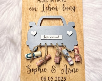 Puede incluir: Un letrero de madera con un recorte de coche gris, las palabras "Hand in Hand ein Leben lang" y "Just married" en él. El coche tiene billetes enrollados atados con cuerda. Los nombres "Sophie & Arne" y la fecha "08.05.2025" también están en el letrero.