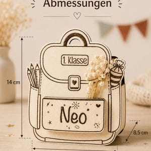 Puede incluir: Decoraci&oacute;n de madera con forma de mochila, de 14 cm de alto, 12 cm de ancho y 8,5 cm de profundidad, con el nombre "Neo" y el texto "1. Klasse". La decoraci&oacute;n incluye detalles de l&aacute;pices y flores.