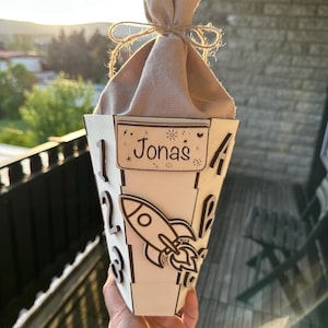 Puede incluir: Una caja de regalo de madera personalizada en forma de cono con el nombre "Jonas" en un peque&ntilde;o letrero rectangular. La caja presenta un dise&ntilde;o de cohete cortado con l&aacute;ser y n&uacute;meros en el lateral. La parte superior est&aacute; cubierta con tela beige y atada con hilo.