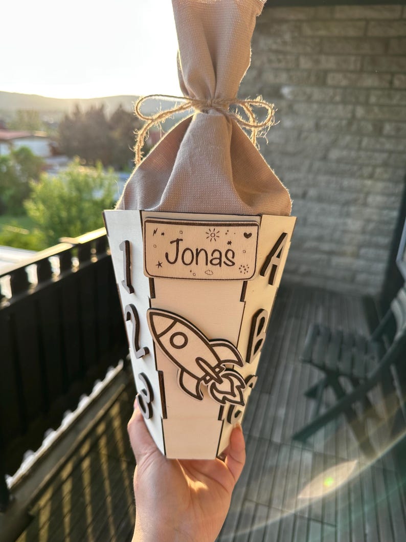 Puede incluir: Una caja de regalo de madera personalizada en forma de cono con el nombre "Jonas" en un peque&ntilde;o letrero rectangular. La caja presenta un dise&ntilde;o de cohete cortado con l&aacute;ser y n&uacute;meros en el lateral. La parte superior est&aacute; cubierta con tela beige y atada con hilo.