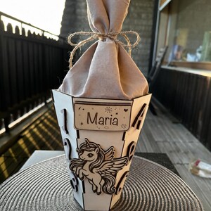 Puede incluir: Una caja de regalo de madera blanca con un dise&ntilde;o de unicornio y el nombre "Maria". La caja est&aacute; rellena de tela beige y atada con hilo. La caja est&aacute; sobre un salvamanteles plateado.