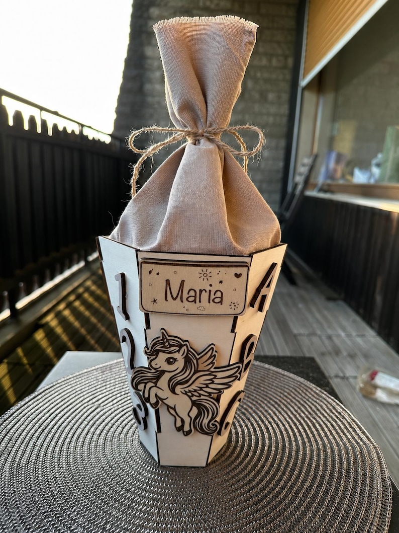 Puede incluir: Una caja de regalo de madera blanca con un dise&ntilde;o de unicornio y el nombre "Maria". La caja est&aacute; rellena de tela beige y atada con hilo. La caja est&aacute; sobre un salvamanteles plateado.