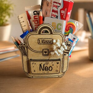 Puede incluir: Un recipiente de madera con forma de mochila lleno de &uacute;tiles escolares y golosinas. El recipiente tiene el texto "1 Klasse" y "Neo". Contenido: l&aacute;pices, una regla, caramelos y una caja de chocolate Kinder.