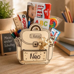 Puede incluir: Un organizador de escritorio de madera con forma de mochila, lleno de &uacute;tiles escolares y golosinas. El organizador tiene el nombre "Neo" en la parte delantera y las palabras "1. Klasse" en la parte superior. Contenido: l&aacute;pices, una regla, caramelos y una caja Kinder Sorpresa.