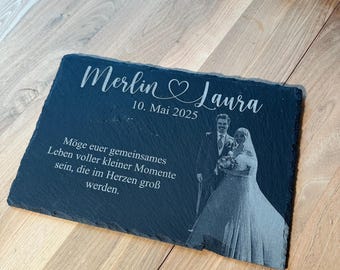 Puede incluir: Una pizarra negra conmemorativa de boda con los nombres "Merlin & Laura" y la fecha "10. Mai 2025" en escritura blanca. Una foto de una pareja está grabada en la pizarra, junto con una cita alemana.