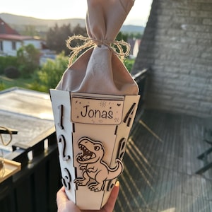 Puede incluir: Un recipiente de madera blanco, en forma de cono, con el nombre "Jonas" en un peque&ntilde;o letrero. Un dinosaurio de dibujos animados est&aacute; en la parte delantera. El recipiente est&aacute; lleno de una bolsa de tela beige atada con hilo. Los n&uacute;meros est&aacute;n recortados del recipiente.