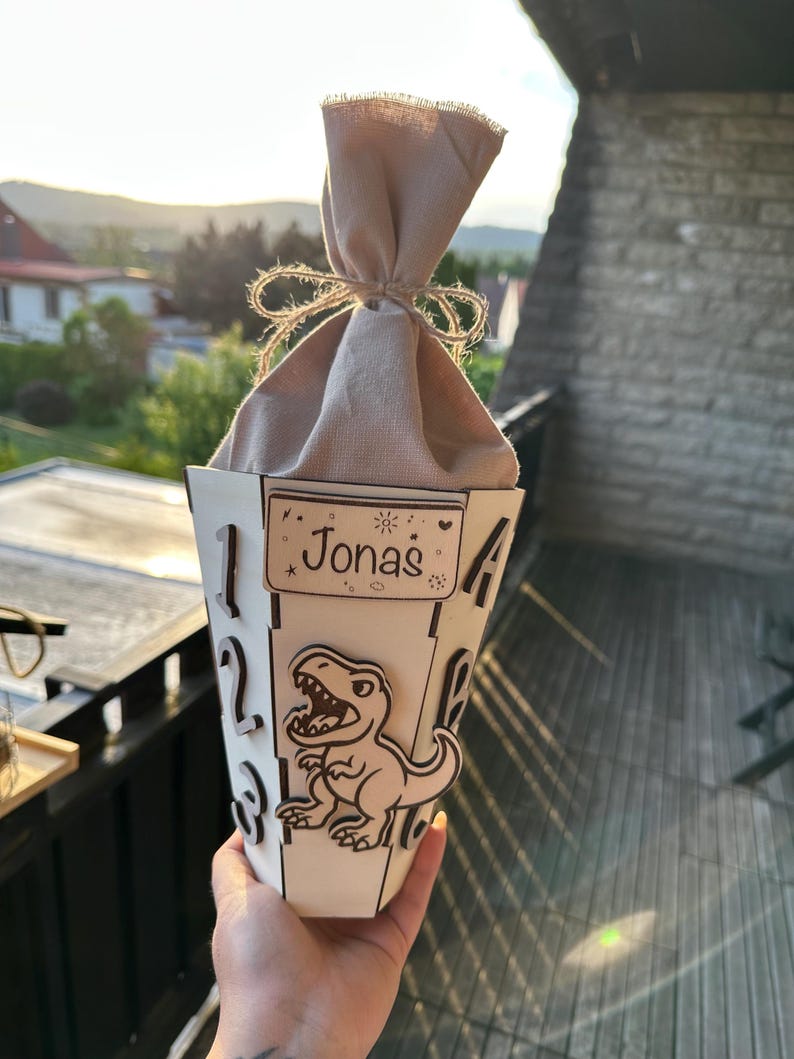 Puede incluir: Un recipiente de madera blanco, en forma de cono, con el nombre "Jonas" en un peque&ntilde;o letrero. Un dinosaurio de dibujos animados est&aacute; en la parte delantera. El recipiente est&aacute; lleno de una bolsa de tela beige atada con hilo. Los n&uacute;meros est&aacute;n recortados del recipiente.