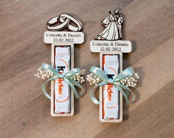 Puede incluir: Dos recuerdos de boda con una barra de chocolate Kinder en un soporte de madera. Cada recuerdo tiene una parte superior de madera con los nombres "Concetta & Dennis" y la fecha "22.02.2022". Decorados con una cinta azul claro y pequeñas flores blancas.