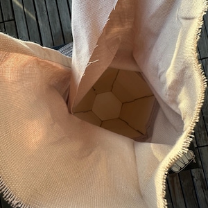 Puede incluir: Una bolsa de arpillera beige con un borde deshilachado, que revela una estructura de madera geom&eacute;trica en el interior. La textura de la bolsa es &aacute;spera y la estructura interior tiene un dise&ntilde;o de panal. La est&eacute;tica general es r&uacute;stica y natural.