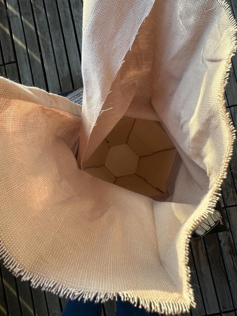 Puede incluir: Una bolsa de arpillera beige con un borde deshilachado, que revela una estructura de madera geom&eacute;trica en el interior. La textura de la bolsa es &aacute;spera y la estructura interior tiene un dise&ntilde;o de panal. La est&eacute;tica general es r&uacute;stica y natural.