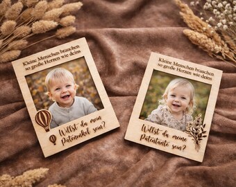 Puede incluir: Dos marcos de fotos de madera con fotos de niños sonrientes. Los marcos tienen texto grabado en alemán, incluyendo "Willst du mein Patenonkel sein?" y "Willst du meine Patentante sein?" Los elementos decorativos incluyen un globo aerostático y detalles florales.
