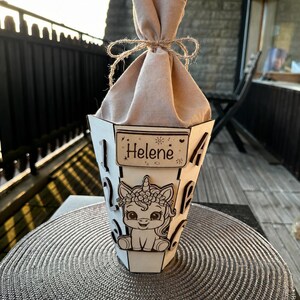 Puede incluir: Un recipiente de madera blanco en forma de cono con un dise&ntilde;o de unicornio y el nombre "Helene" en un peque&ntilde;o letrero. El recipiente est&aacute; lleno de una tela beige atada con hilo.