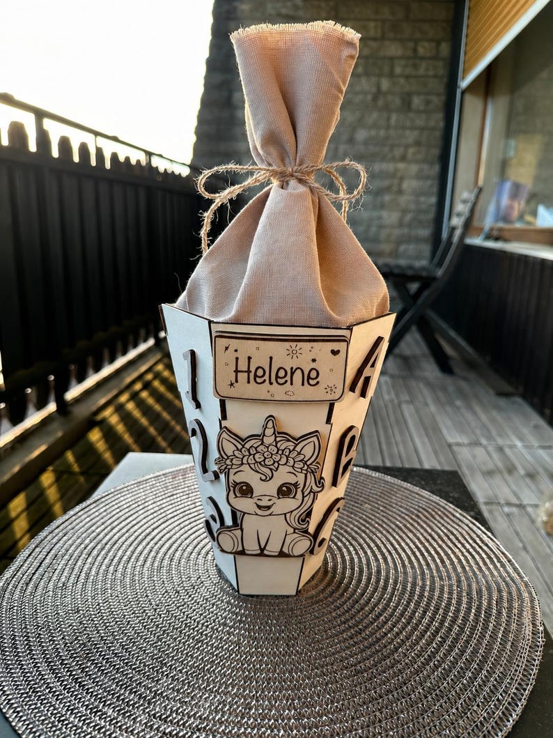 Puede incluir: Un recipiente de madera blanco en forma de cono con un dise&ntilde;o de unicornio y el nombre "Helene" en un peque&ntilde;o letrero. El recipiente est&aacute; lleno de una tela beige atada con hilo.