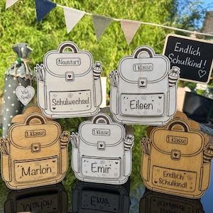 Puede incluir: Decoraciones de madera con forma de mochila con el texto "1. Klasse" y nombres como "Eileen" y "Marlon". Algunas tienen el texto "Endlich Schulkind!" y "Schulwechsel". Tambi&eacute;n se ven l&aacute;pices y una peque&ntilde;a pizarra.