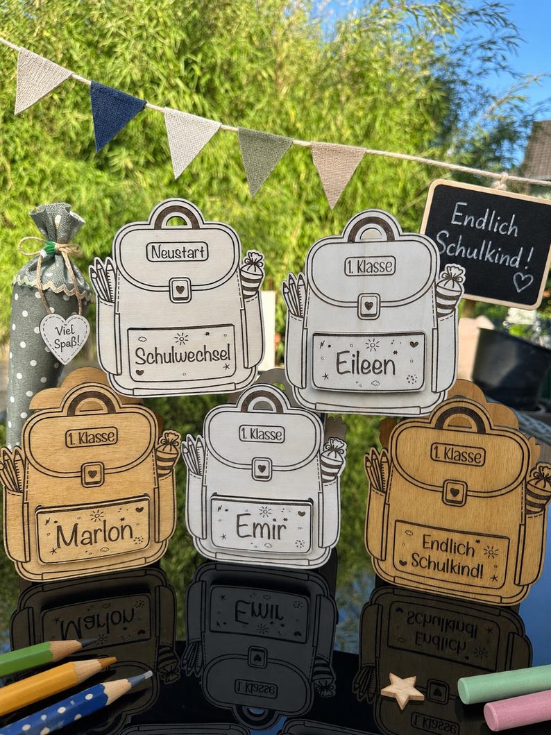 Puede incluir: Decoraciones de madera con forma de mochila con el texto "1. Klasse" y nombres como "Eileen" y "Marlon". Algunas tienen el texto "Endlich Schulkind!" y "Schulwechsel". Tambi&eacute;n se ven l&aacute;pices y una peque&ntilde;a pizarra.