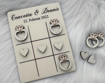Puede incluir: Un juego de tres en raya de madera con los nombres Concetta & Dennis y la fecha 22. Februar 2022. Las piezas del juego son anillos de madera con un diseño de diamante y corazón.