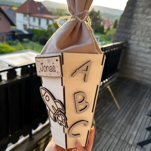 Puede incluir: Una caja de regalo de madera con el nombre "Jonas" y las letras "ABC" recortadas. Tambi&eacute;n hay un cohete recortado en el lateral. La caja est&aacute; rematada con una bolsa de tela beige atada con hilo.