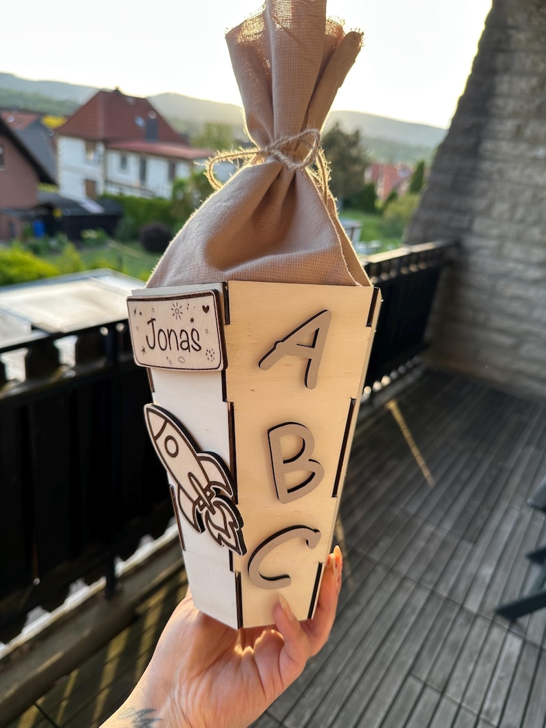 Puede incluir: Una caja de regalo de madera con el nombre "Jonas" y las letras "ABC" recortadas. Tambi&eacute;n hay un cohete recortado en el lateral. La caja est&aacute; rematada con una bolsa de tela beige atada con hilo.