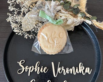 Puede incluir: Un plato negro contiene una galleta en un envoltorio transparente atada con una cinta verde y un ramo de flores secas. La galleta está grabada con iniciales y la fecha 08.10.2022. Los nombres "Sophie Veronika Gerlinde" se muestran.