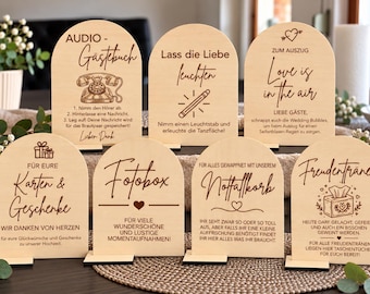 Puede incluir: Siete letreros de madera clara en forma de arco con texto e iconos grabados en marrón oscuro. Los letreros presentan frases en alemán que incluyen Audio-Gästebuch, Lass die Liebe leuchten, Love is in the air, Karten & Geschenke, Fotobox, Notfallkorb y Freudentränen.