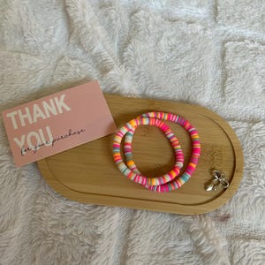 May include: Two colourful beaded bracelets with pink, orange, yellow, and blue accents sit on a wooden tray. A pink card with the words "THANK YOU for your purchase" is also on the tray, along with a silver charm. The bracelets are on a white, fluffy surface.
