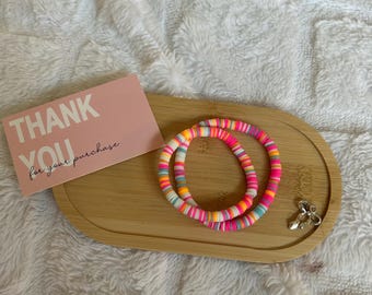 Op de afbeelding: Twee kleurrijke kralenarmbanden met roze, oranje, gele en blauwe accenten liggen op een houten dienblad. Een roze kaart met de tekst "THANK YOU for your purchase" ligt ook op de dienblad, samen met een zilveren bedel. De armbanden liggen op een witte, zachte ondergrond.