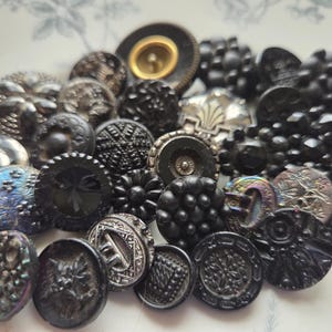 May include: A collection of assorted black buttons in various shapes and sizes. The buttons have different textures, including smooth, ridged, and patterned designs. Some buttons have iridescent or metallic finishes, and some have gold or silver accents.