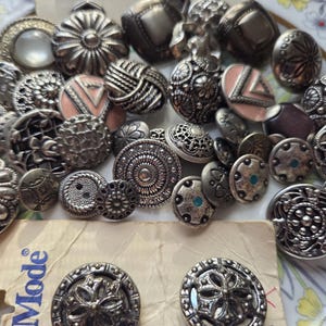 May include: A collection of assorted metal buttons in various circular shapes and intricate embossed patterns, featuring silver-toned finishes, some with pink or blue accents, alongside two matching buttons attached to a beige Mode brand card.