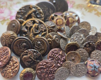 May include: A selection of antique buttons in various sizes and designs. The buttons are mainly bronze, copper, and silver in colour, with intricate embossed patterns, including floral, figural, and textured details. Some buttons have a deep red centre.
