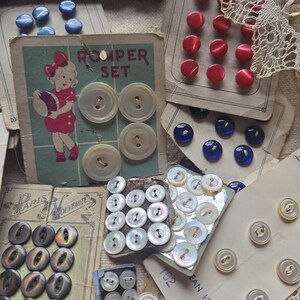 May include: A collection of vintage buttons in various colors and sizes. Includes red, blue, and iridescent mother-of-pearl buttons, some still attached to their original cards. A card with a child illustration and the words "Romper Set" is also visible.