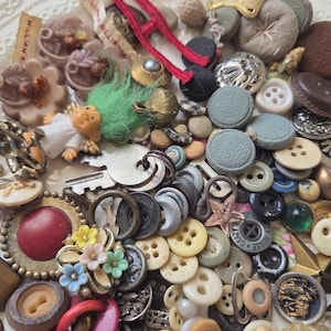 May include: A collection of vintage buttons and embellishments in various shapes, sizes, and colors. Includes fabric-covered, metal, and plastic buttons, along with small figurines, keys, and brooches. The color palette includes brown, blue, green, and cream.