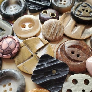 May include: A collection of vintage buttons in various colors, shapes, and materials, including brown, black, white, pink, and beige. The buttons are arranged on a white plate with a floral pattern.